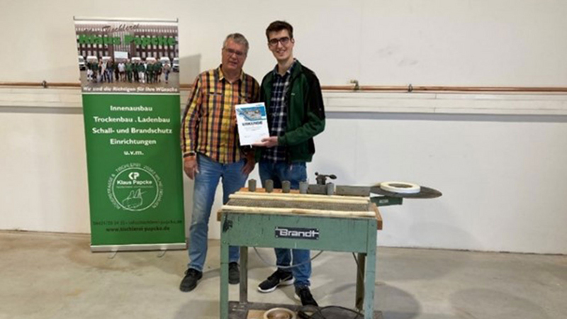 公司创始人 Klaus Papcke 和他的儿子 Phil 正在展示获奖的台式封边机 T-50，这是 1978 年生产的 BRANDT 机型